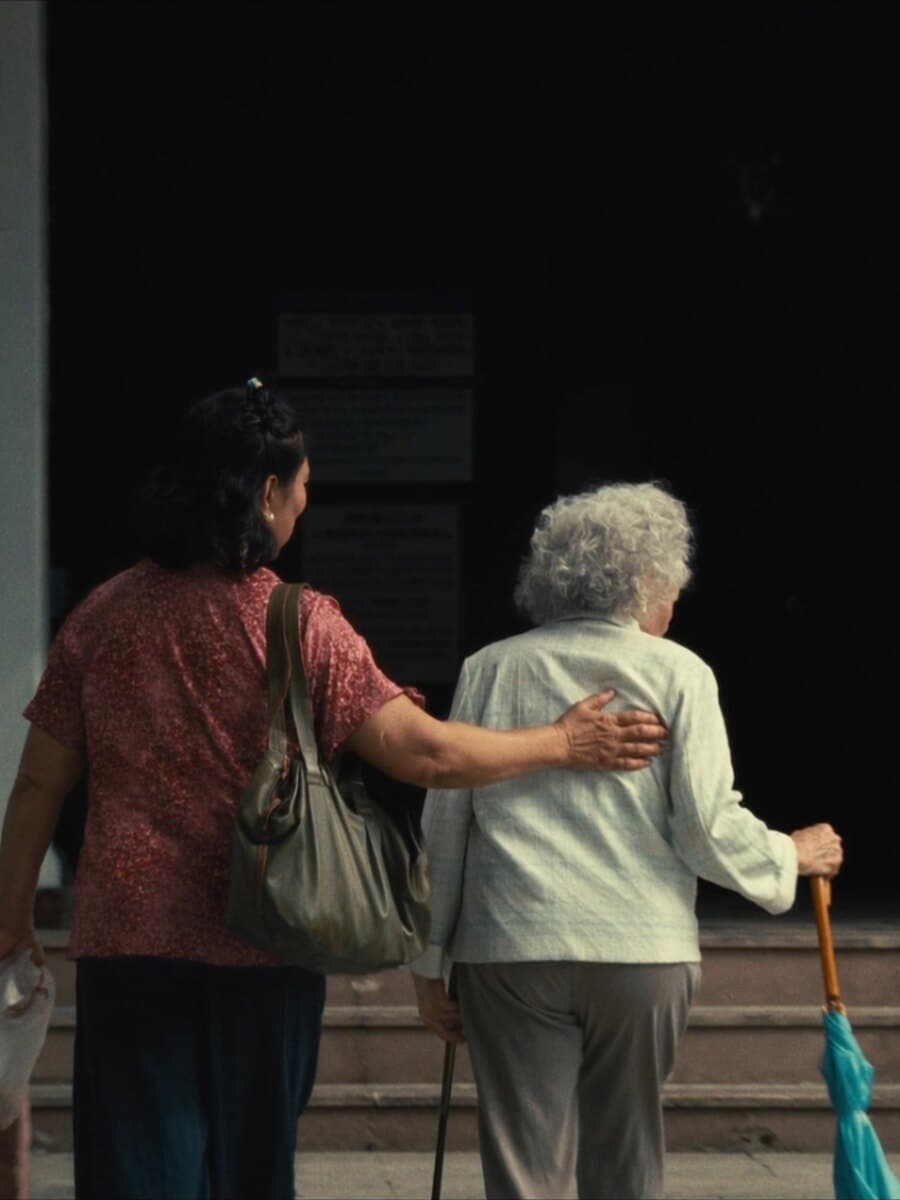 A caregiver walks arm in arm with an elderly woman toward a clinic entrance